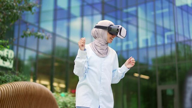 Happy young muslim female student in hijab dancing using VR glasses on the street near modern university building. Crazy joyful girl having fun in virtual reality simulator. Dance of a funny teenager