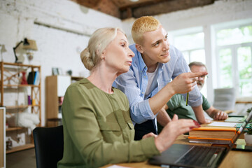 Fototapeta premium Young man, employee heping senior worker, colleges with professional issues, teaching to use laptop apps for presentation. Concept of business, diversity, education, modernity, innovations, assistance
