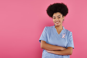 smiling african american doctor with ribbon crossing arms isolated on pink, breast cancer concept