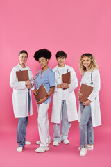 Fototapeta premium smiling multiethnic doctors with paper folders standing together on pink, breast cancer awareness