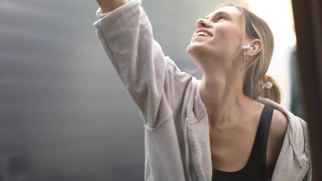 Young smiling woman washing window with green sponge. Happy beautiful girl cleaning window by spraying cleaning products and wiping with sponge in yellow gloves, wireless earphones, dance, sing song.