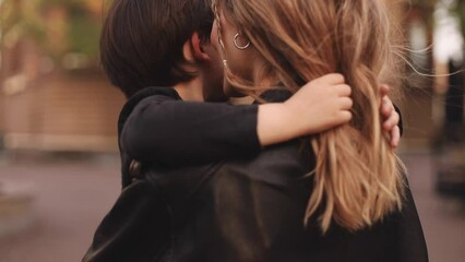 Attractive blonde curly woman with two thin braids wear black leather jacket and dress carries in her arms her brunette son, he hugs mothers neck, and touch her hairs, family go home. Selective focus.