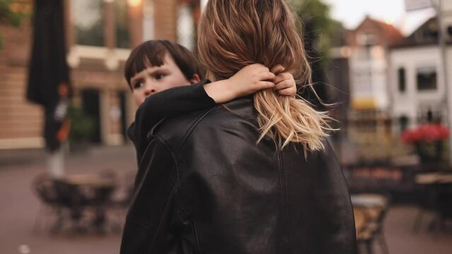 Attractive blonde curly woman with two thin braids wear black leather jacket and dress carries in her arms her brunette son, he hugs mothers neck, and touch her hairs, family go home. Boy tired walk.