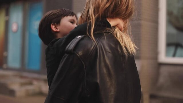 Attractive blonde curly woman with two thin braids wear black leather jacket and dress carries in her arms her brunette son, he hugs mothers neck, and touch her hairs, family go home. Selective focus.