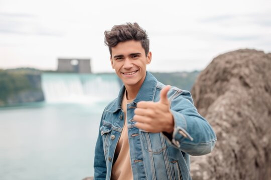 Headshot portrait photography of a grinning boy in his 20s making a stop sign with hand sporting a rugged denim jacket at the niagara falls in ontario canada. With generative AI technology