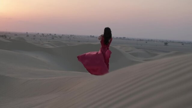 Young woman with black hair walking barefoot in a long red evening dress on the sand dunes of the desert