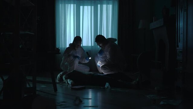 The forensic team examines the body of the victim, a murdered woman, and collects evidence using a flashlight and tongs. Specialists in a dark apartment, lit by red and blue police sirens.