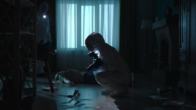 A team of forensic scientists collect evidence at the crime scene, in a dark apartment lit by blue red light. A man inspects a crime scene using a flashlight. A woman photographs evidence.