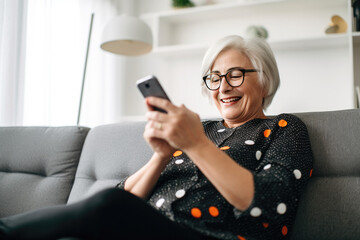 Modern lifestyle of the elderly. Stylish senior woman talking online using smartphone while relaxing on sofa. - obrazy, fototapety, plakaty