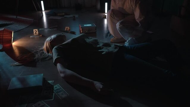 The forensic expert leans over the woman's body and uses an ultraviolet lamp to look for traces and DNA samples. A criminologist works at a crime scene in a dark apartment lit by blue red light.