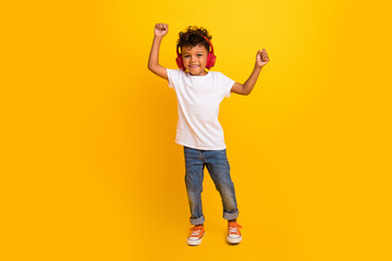 Full length photo of positive adorable small boy wear white t-shirt having fun listening music headphones isolated yellow color background