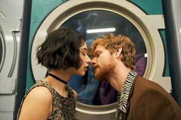 young and stylish multiethnic couple kissing near washing machine in coin laundry