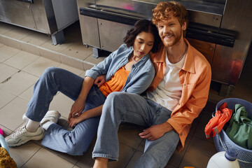 high angle view of smiling multiethnic couple sitting near washing machine in coin laundry