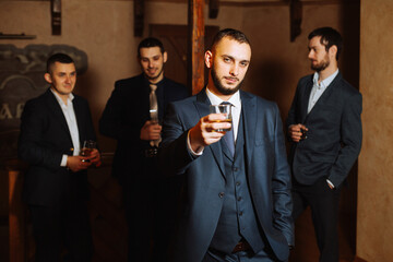 the groom in the foreground and his friends in stylish suits drinking whiskey in the hotel room, the morning before the wedding preparations