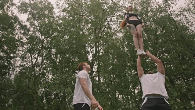 Low angle shot of strong male athlete tossing and holding cheerleader flyer above his head while practicing extension stunt outdoors