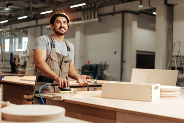 Young carpenter sanding wood piece in workshop in furniture factory