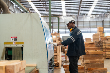 Fototapeta premium Black man worker operating CNC machine for cutting wood in factory. Male engineer in uniform with helmet safety working produce wooden pallet at manufacturing plant.