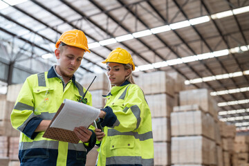 Man and woman workers in uniform with helmet safety holding clipboard discussing and checking wood plank material in pallet factory warehouse.
