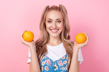 Portrait of toothy beaming pretty girl with ponytails wear t-shirt under dress arms hold two oranges isolated on pink color background