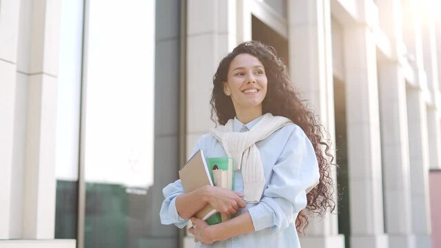 A happy smiling female student walks down the street in the campus space near the university building. Positive curly brunette girl with books, textbooks in her hands goes to a lecture at the college