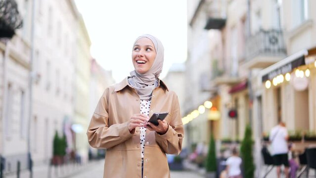 Attractive young Muslim young woman tourist in hijab walking on city street using smartphone mobile phone Outdoors browsing internet, searching. Technology lifestyle travel trip and journey concept