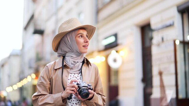 Happy muslim young female tourist in a hat walking along the city street. Lovely lady enjoy the day street lifestyle a girl takes a photo on a camera having good time wandering around town in summer