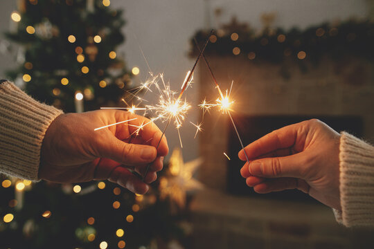 Hands holding burning fireworks against modern fireplace and christmas tree with golden lights. Happy New Year! Friends and family celebrating with burning sparklers in hands, atmospheric eve