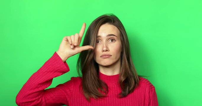 You lose! Brunette young woman making loser gesture, L sign on forehead, teasing and accusing for defeat, expressing disrespect. Indoor studio shot isolated on green background.