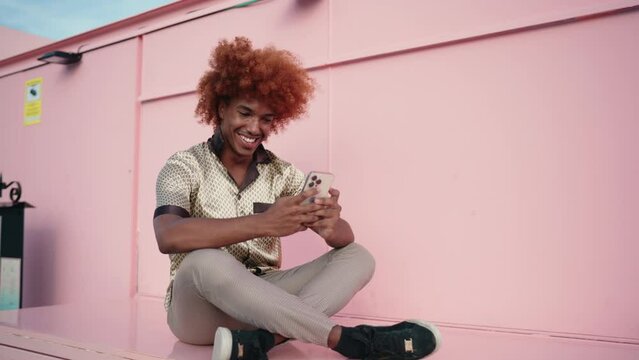 Cheerful African American man with curly hair sits on vibrant pink copy space background, engrossed in smartphone app, sending messages with smile. Happy guy enjoying internet connection on cellula