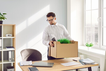 Fired male corporate employee packing his stuff at former workplace. Now unemployed young man in white shirt standing by office work desk and putting his belongings in cardboard box. Job loss concept