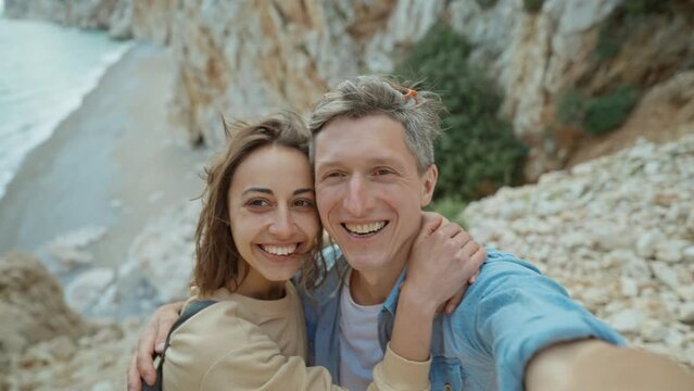 happy lovely couple young smiling woman and mid aged man hiking rocky canyon at seaside and taking selfie. wanderlust and travel summer vacation trip. using phone camera for filming