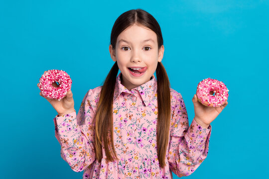 Photo portrait of cute little girl hold two dougnuts lick lips dressed stylish pink flower print clothes isolated on blue color background