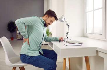 Tired young office worker suffering from back pain while working on laptop at the desk. Attractive...
