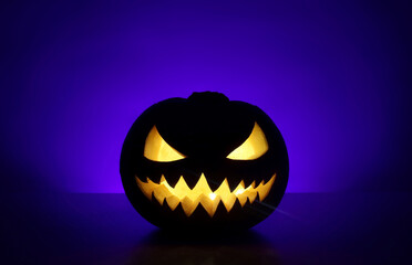 Halloween holiday background with traditional spooky angry jack-o-lantern. Carved glowing pumpkin stands on surface in dark night. Back wall with place for text is highlighted blue with shadows.