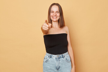 Smiling cheerful brown haired young woman wearing black top and jeans standing isolated over beige background pointing to camera choosing you selecting
