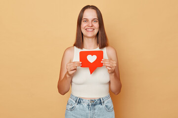 Blogger recommends content. Clicking the button. Social media networking. Smiling happy brown haired young woman wearing casual clothing holding red like icon standing isolated over beige background