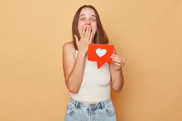 Blog post comments. Web forum engagement. Follow for updates. Surprised brown haired young woman wearing casual clothing holding red like icon standing isolated over beige background