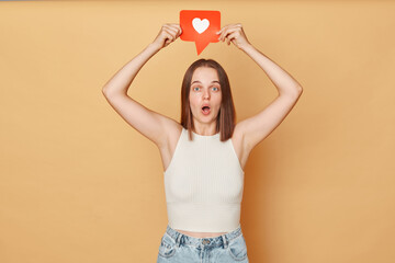 Blogging communication. Heart icon click. Online community interaction. Surprised excited brown haired young woman wearing casual clothing holding red like icon standing isolated over beige background