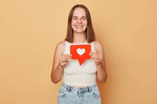 Blogger recommends content. Clicking the button. Social media networking. Smiling happy brown haired young woman wearing casual clothing holding red like icon standing isolated over beige background