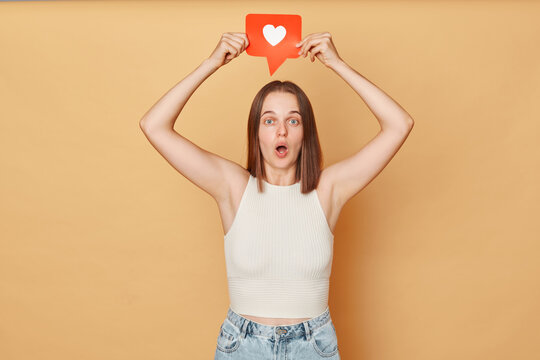 Blogging communication. Heart icon click. Online community interaction. Surprised excited brown haired young woman wearing casual clothing holding red like icon standing isolated over beige background