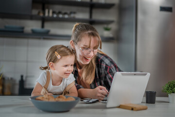 One young woman with toddler girl in the kitchen making a video call and playing with finger...