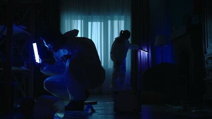A team of forensic specialists work at the crime scene, in a dark apartment lit by blue light from...