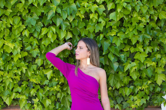 Pretty young blonde woman in purple dress posing for photos against background of a wall of ivy. Woman makes different expressions, serious, happy.