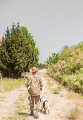 Unrecognizable male fisher in straw hat and camouflage walking down country road with spotted...