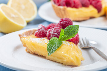 Piece of lemon tart on white plate, garnished with fresh raspberry, icing sugar, mint, horizontal