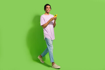 Full length photo of funny charming man dressed purple t-shirt texting modern device walking empty space isolated green color background