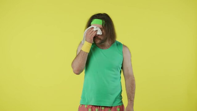 Man in green top and headband standing, wiping his sweating face with white towel. Isolated on yellow background.