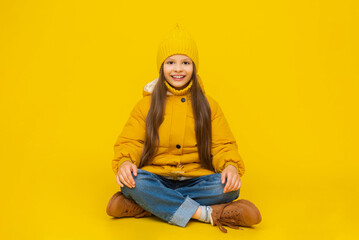 A young girl is sitting on the floor in full height, cross-legged, in autumn warm clothes. A child...