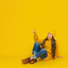 A young girl is sitting on the floor in full height, cross-legged, in autumn warm clothes. A child in a yellow warm jacket and a cap points his finger up.