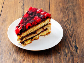 Slice of tasty multi layered cake with raspberry and blue berry in jelly on top. Portion of delicious dessert on white plate and wooden table. High quality bakery product serving in coffee shop.